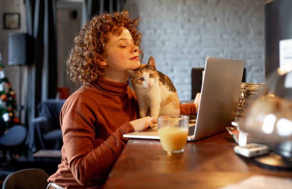 A person with curly hair sits at a desk using a laptop, smiling and leaning close to a cat that is sitting beside the laptop. A glass of orange juice is on the desk in the foreground.