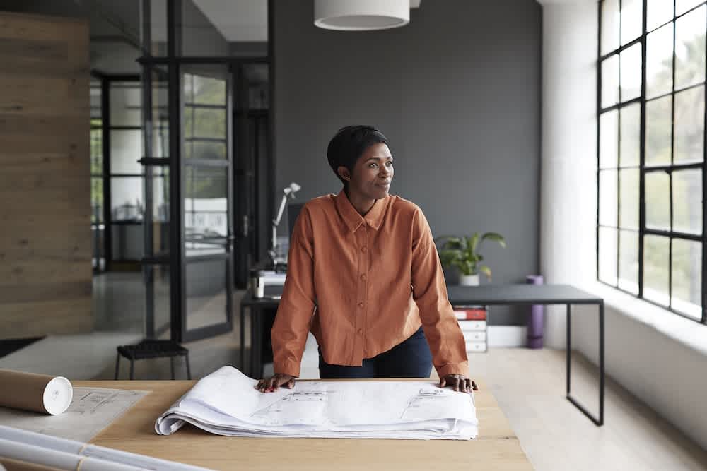 A woman wearing an orange shirt stands at a table with architectural plans, looking out a large window in a modern office space filled with natural light.