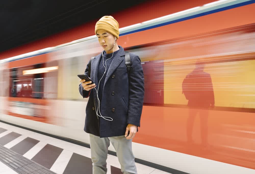 A person wearing a yellow beanie, dark coat, and backpack stands on a subway platform, looking at their phone with earphones in, while a red train passes by in the background.