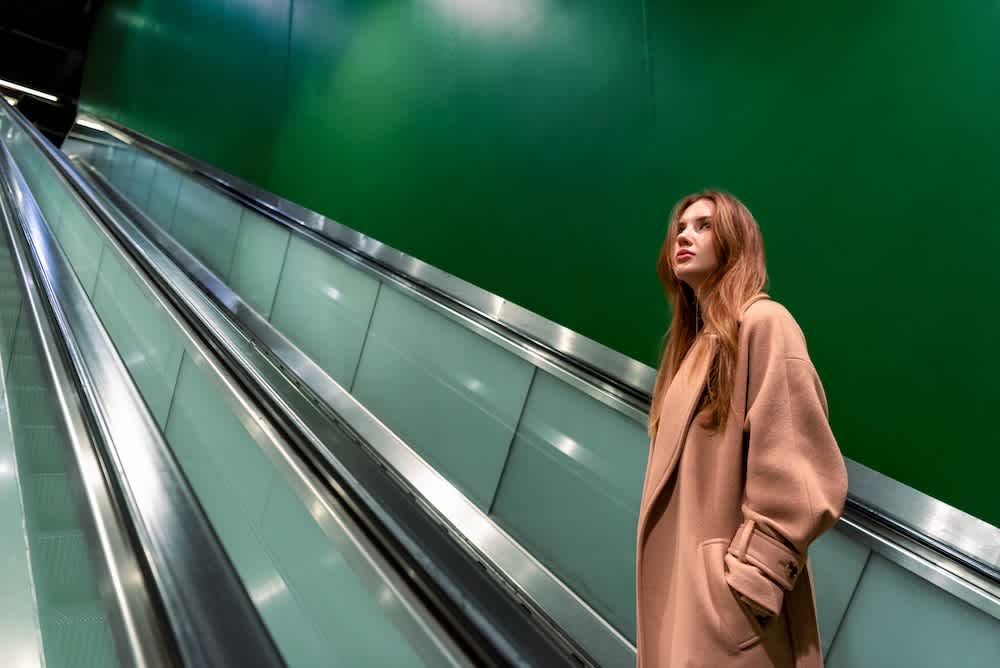 A woman in a tan coat stands beside a modern escalator with metallic sides and a green wall background, looking slightly upward with a thoughtful expression.