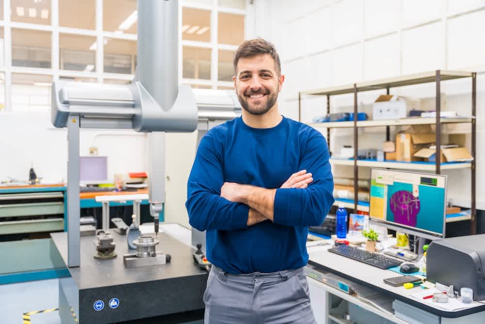A man with a beard and blue shirt stands smiling with arms crossed in a modern laboratory or engineering workspace, with machines, computers, and shelves in the background.