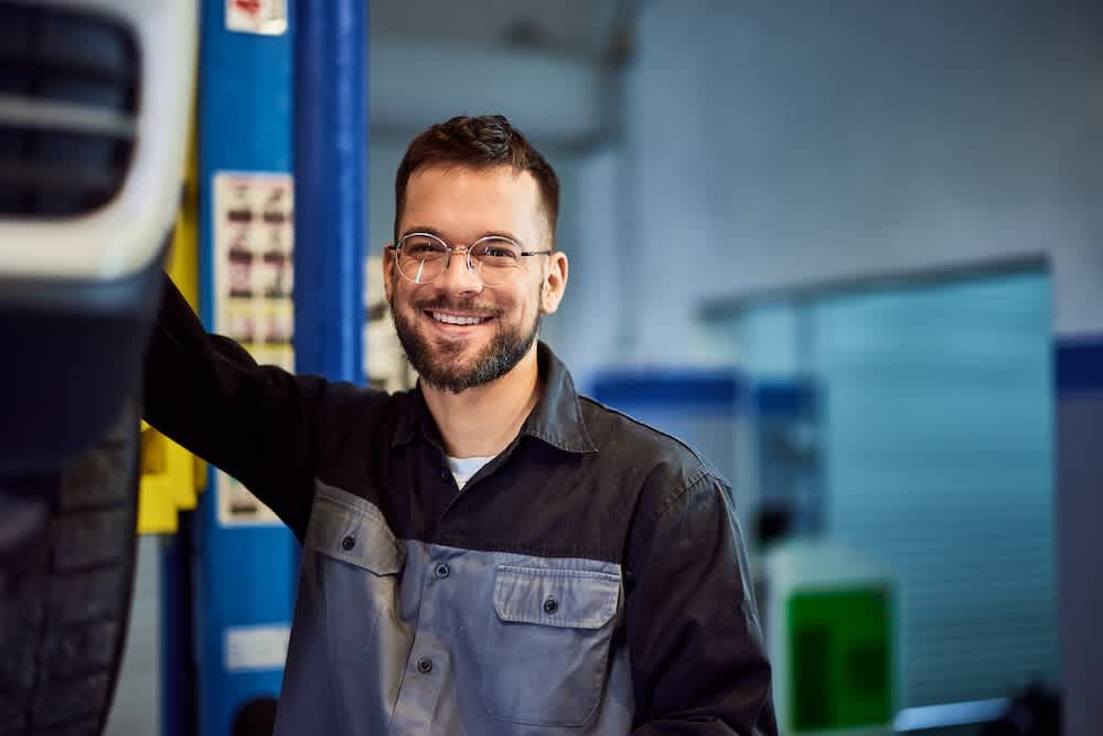 A smiling auto mechanic wearing glasses and a work uniform stands beside a vehicle in a brightly lit garage.