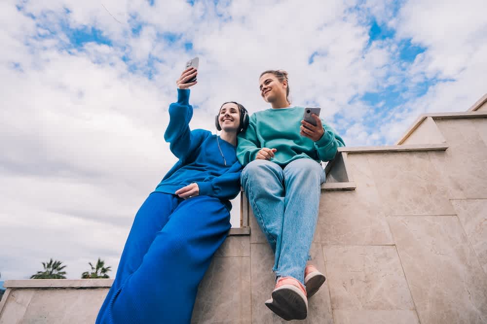 Two young people sit on outdoor steps, smiling and holding smartphones. One is taking a selfie while the other looks at them. The sky is partly cloudy, and both wear casual blue outfits.