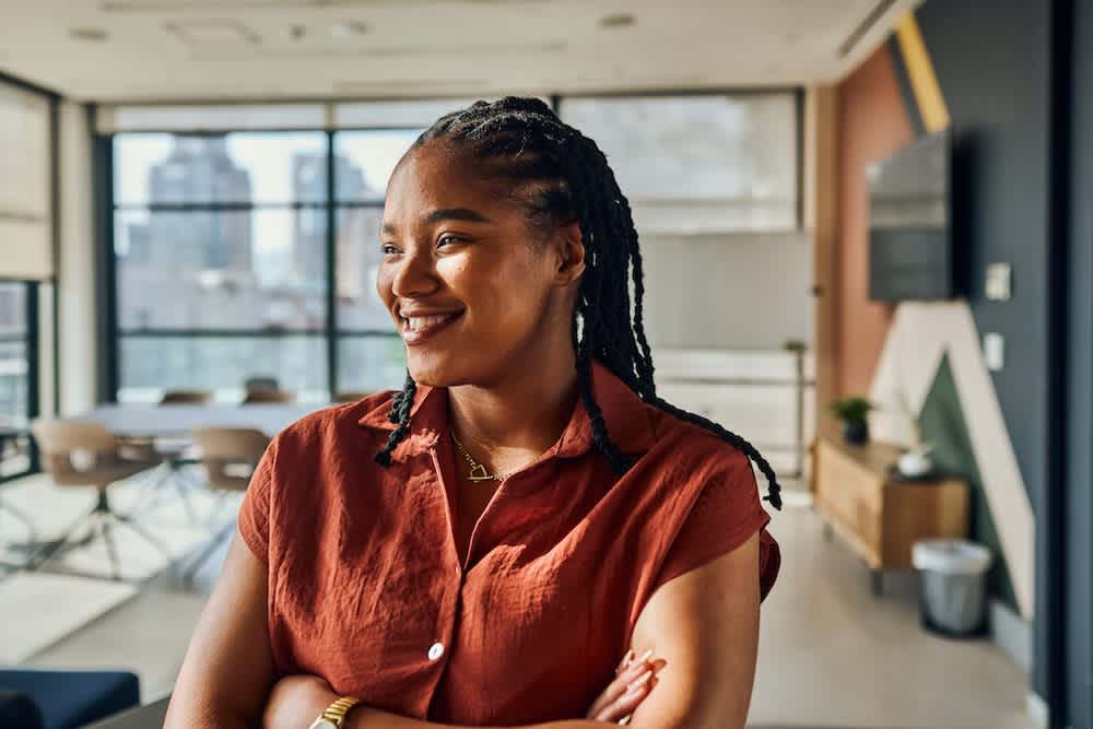 A woman with braided hair, wearing a short-sleeved rust-colored shirt, smiles while standing with arms crossed in a modern, sunlit office space with large windows and city views.
