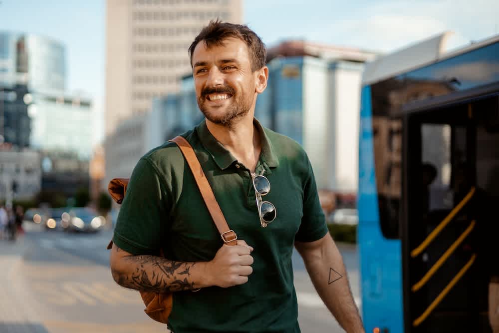 A smiling man with a mustache, wearing a green polo shirt and carrying a brown backpack, stands outdoors near a blue bus. He has tattoos on his arm and sunglasses hanging from his shirt. City buildings are visible in the background.