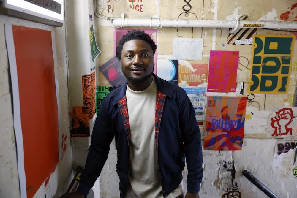 A man stands indoors by a staircase, smiling at the camera. The wall behind him is covered with colorful, eclectic posters and graffiti-style art.