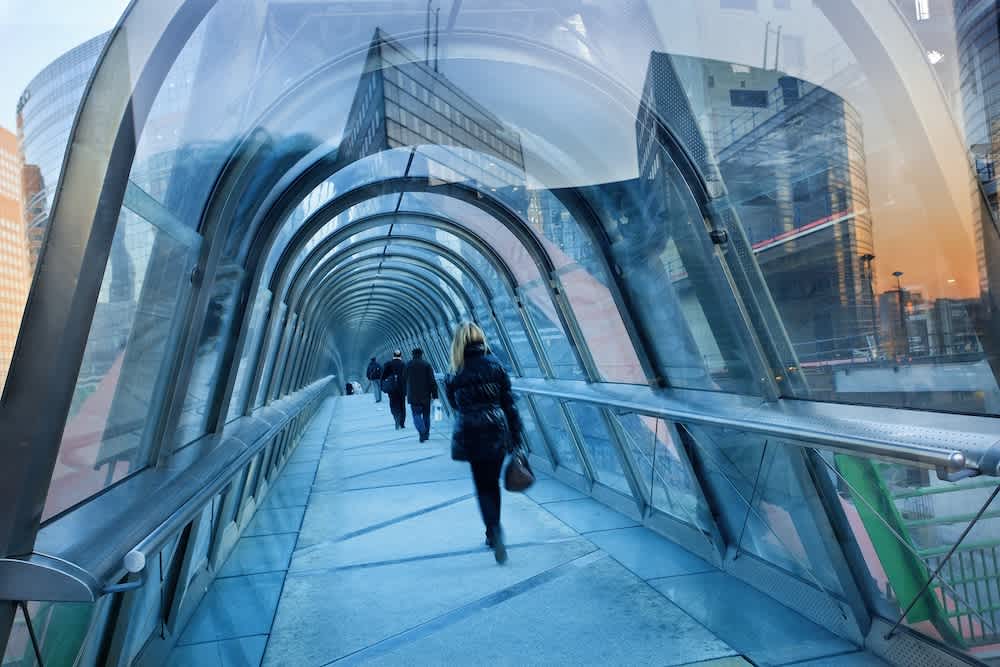 People walk through a modern, glass-covered pedestrian walkway in an urban area, with reflections of city buildings visible on the transparent walls.