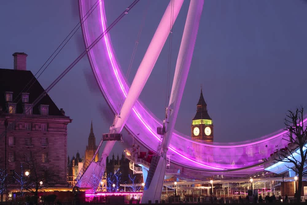 The London Eye is illuminated with purple lights at dusk, creating a blurred arc. Big Ben and the Houses of Parliament are visible in the background, with a clock face glowing against the evening sky.