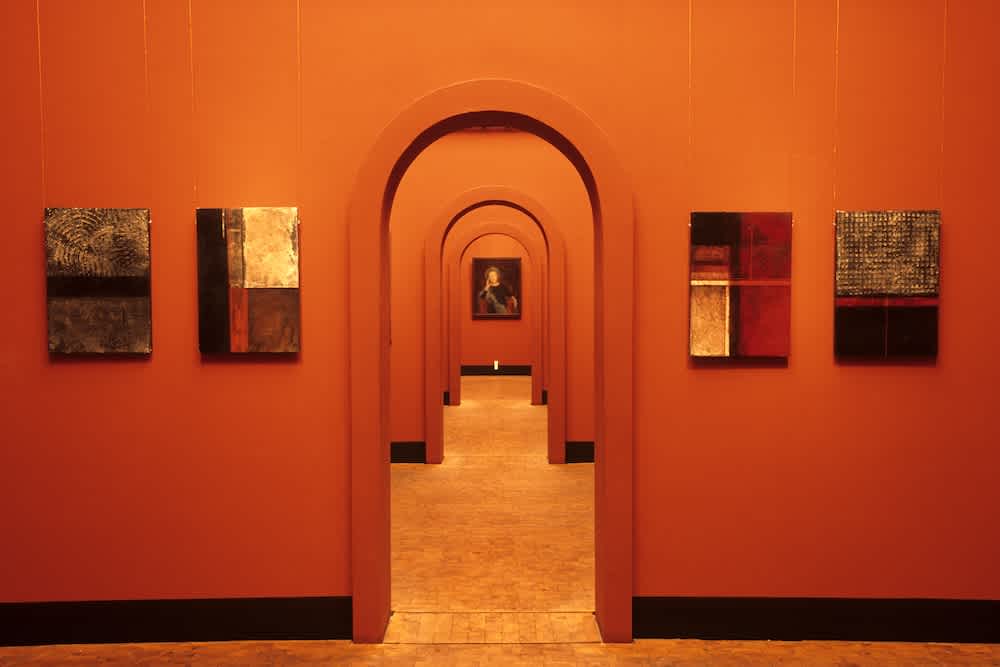 A gallery with terracotta-colored walls features modern, abstract paintings. A series of arched doorways create a tunnel effect, leading to a classical portrait at the far end. The floor is wooden.