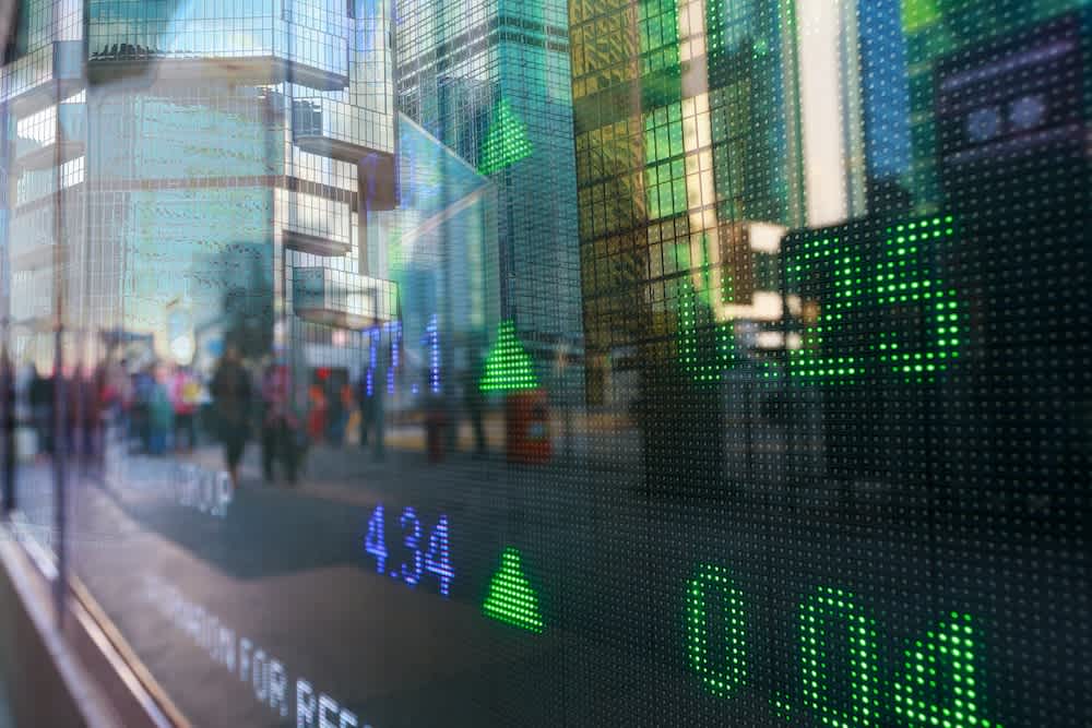 A glass wall displays green digital numbers and arrows indicating rising stock prices, with a city street and pedestrians reflected in the background.