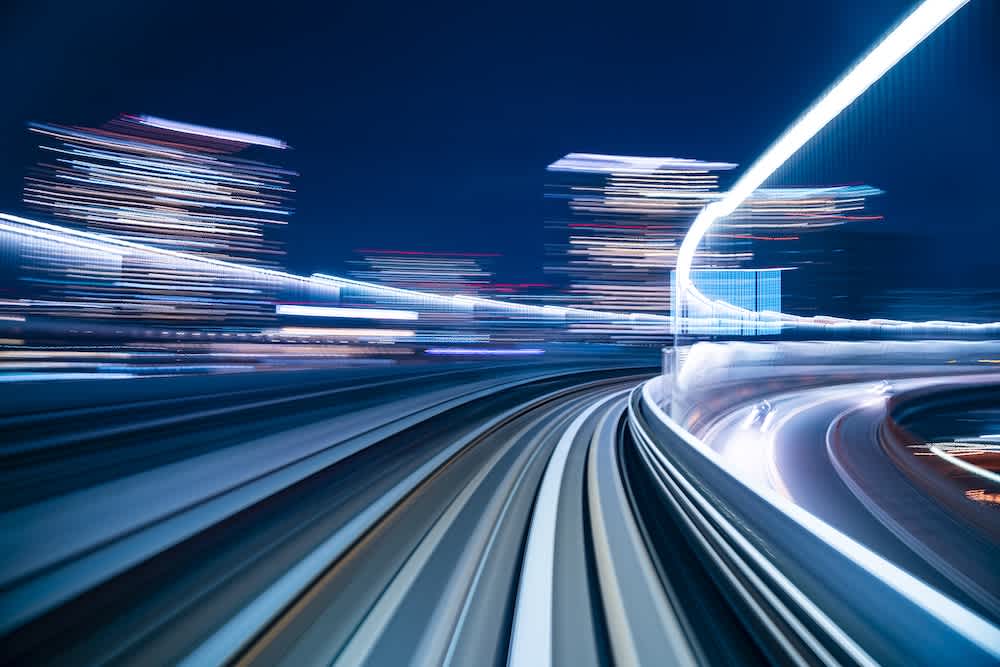 Long-exposure photo of a cityscape at night, showing blurred lights and motion on elevated railway or road tracks, creating a sense of speed and movement through an urban environment.
