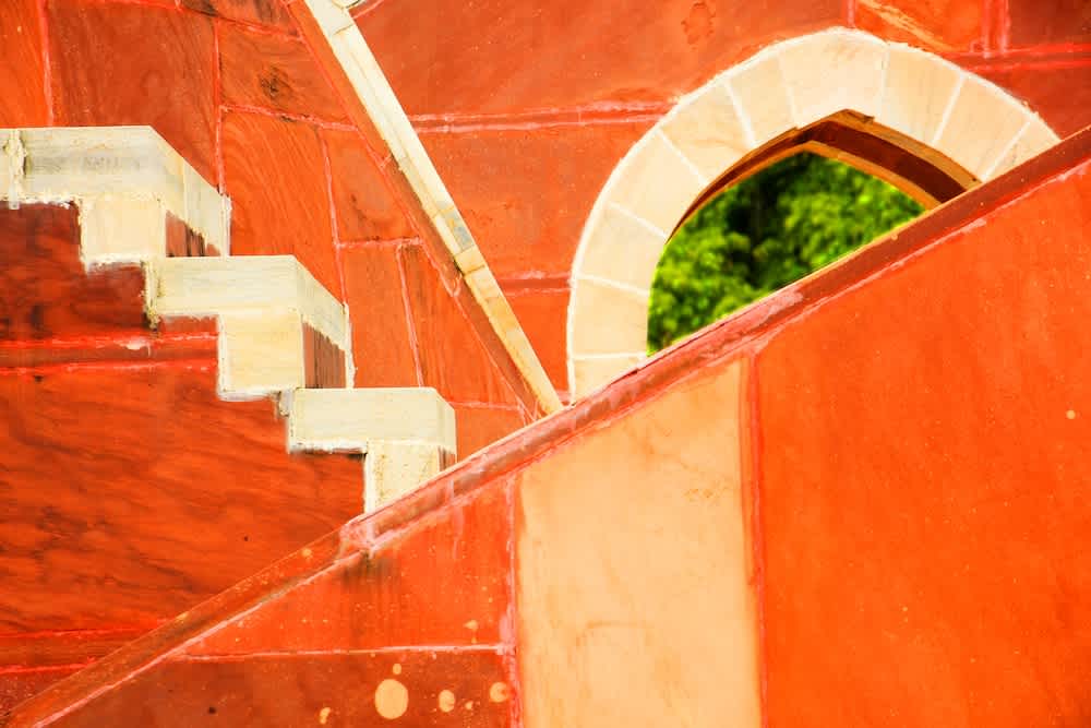 Abstract view of a red sandstone and cream-colored stone staircase, an arched doorway, and geometric walls, with a glimpse of green foliage visible through the arch.