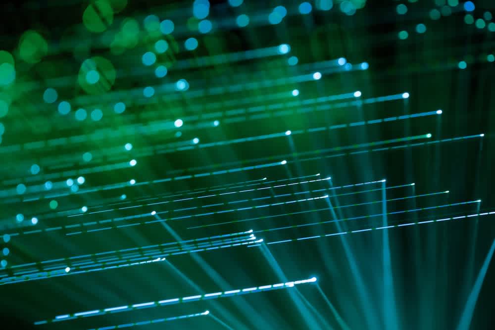 Green and blue fiber optic cables glowing in the dark, creating a dynamic pattern of light streaks and dots against a blurred background.