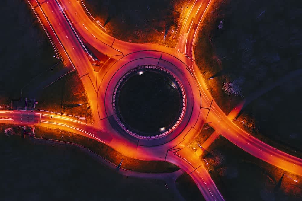Aerial view of a roundabout at night, illuminated by orange streetlights, with multiple roads converging and cars headlights creating bright light trails around the circular intersection.