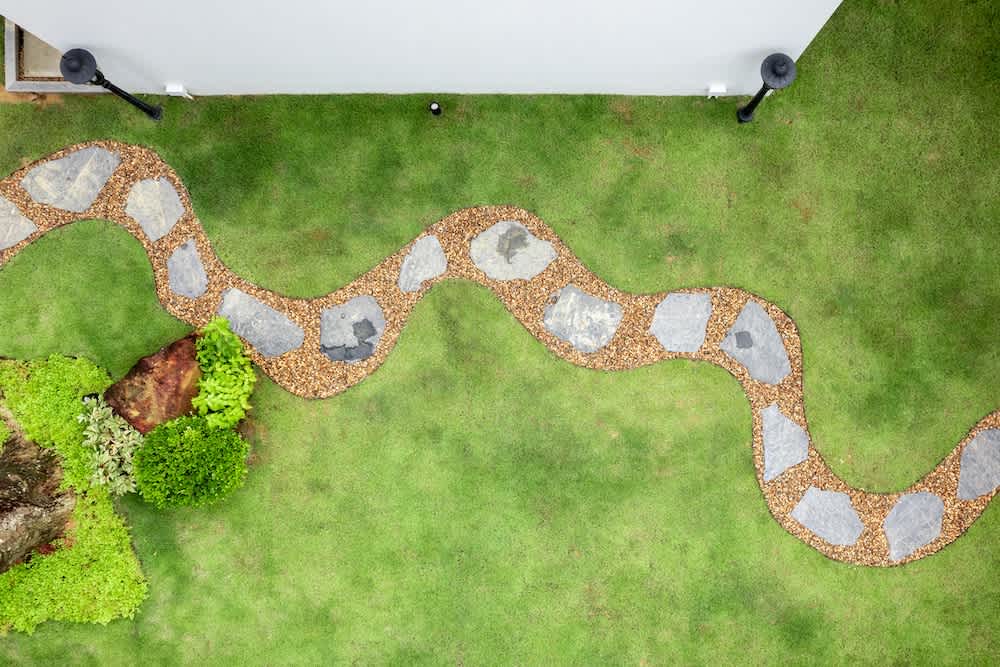 A winding stone path bordered by small pebbles runs across a neatly trimmed green lawn, with greenery and a large rock on the left side, viewed from above.
