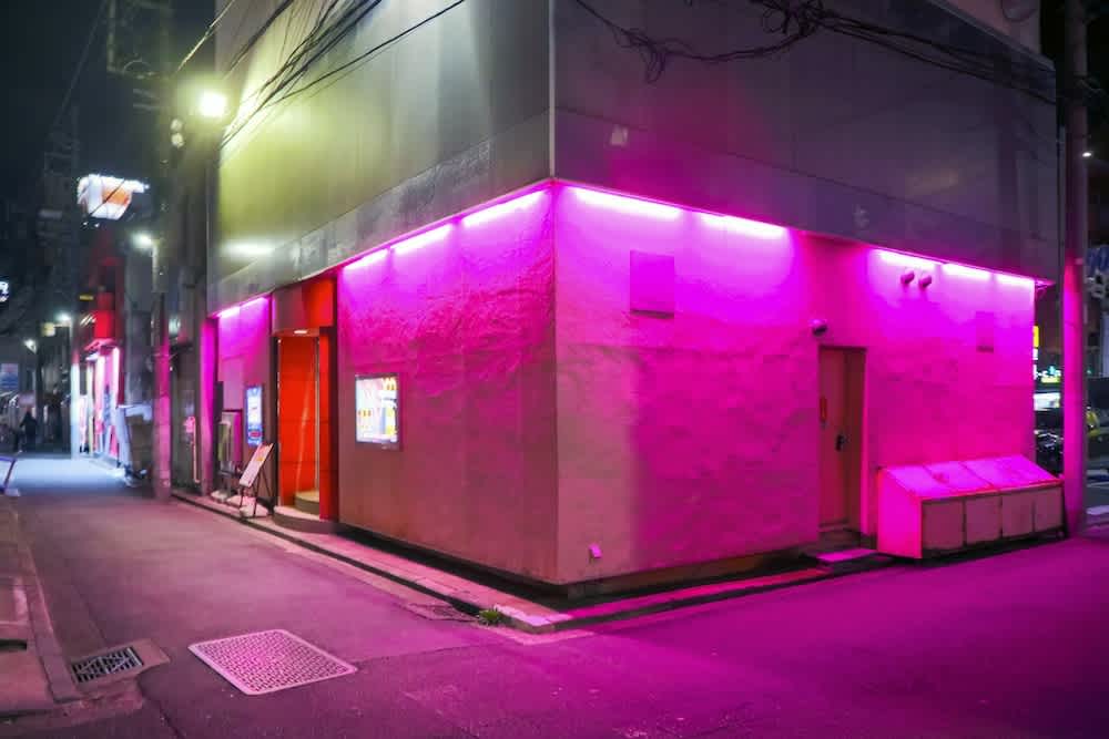 A building on a city street corner is illuminated by vibrant pink and purple neon lights at night, casting a colorful glow on the sidewalk and surrounding area.
