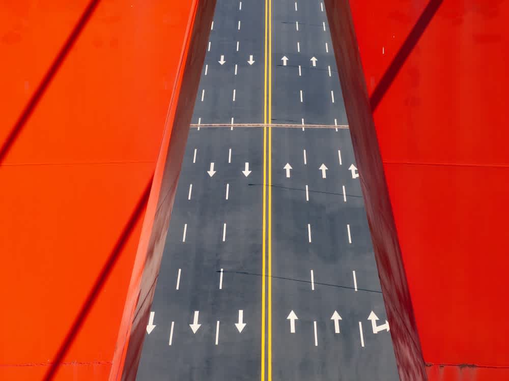 A symmetrical aerial view of a wide empty road with multiple white arrows, flanked by bright red walls or barriers casting shadows. The scene creates a bold geometric and abstract composition.