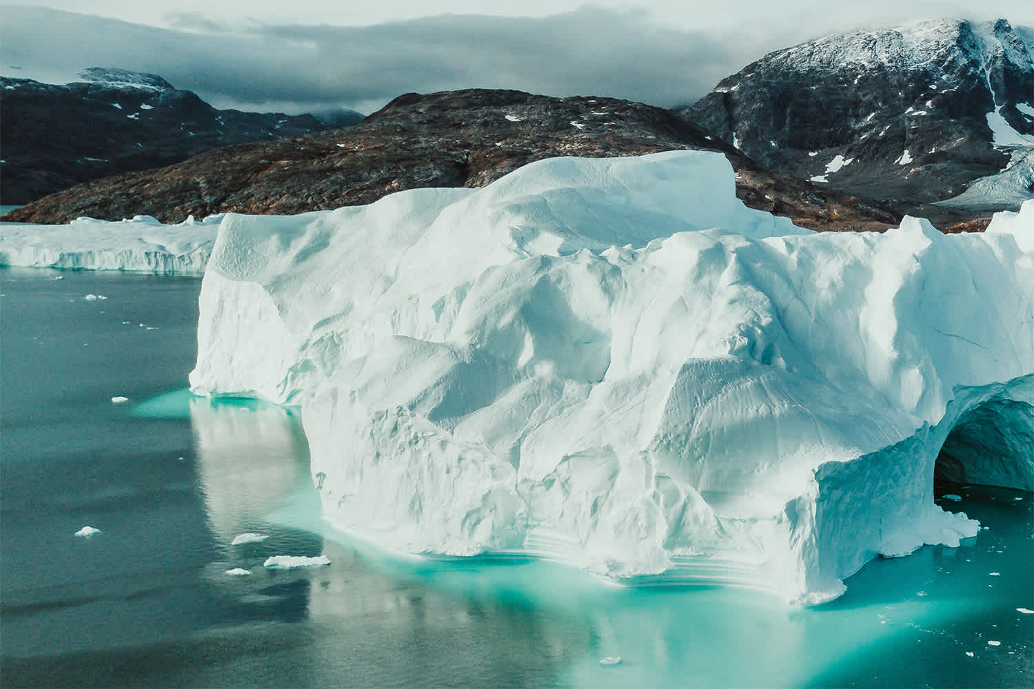 Large white iceberg floats in turquoise water near a rocky, snow-capped shoreline under a cloudy sky, showcasing contrasting textures and colors in a cold, arctic landscape.