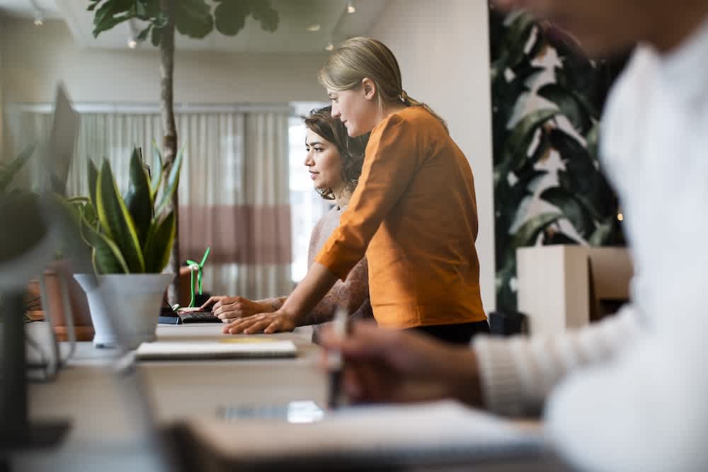 Two women standing at a desk and looking at a computer monitor in a modern office, with a plant and notebooks nearby. A third person is blurred in the foreground, writing in a notebook.