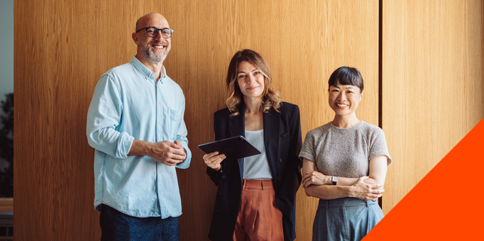 Three people stand smiling in front of a wooden wall. The man on the left wears glasses and a light blue shirt. The woman in the middle holds a tablet, and the woman on the right stands with arms crossed. An orange triangle is in the bottom right corner.