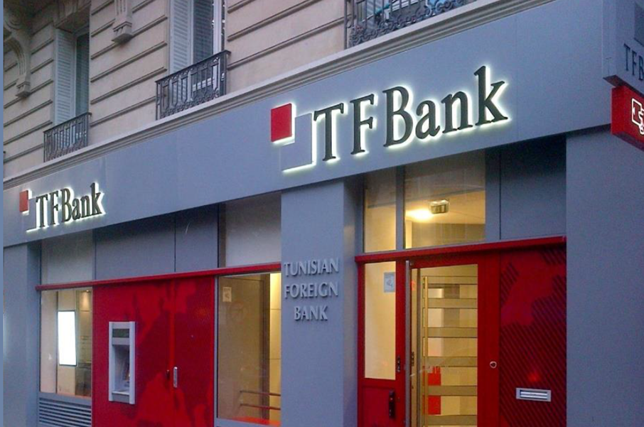The exterior of a TF Bank branch, with large illuminated signs and a glass entrance door. The building facade is gray with red accents, and windows display the text Tunisian Foreign Bank.