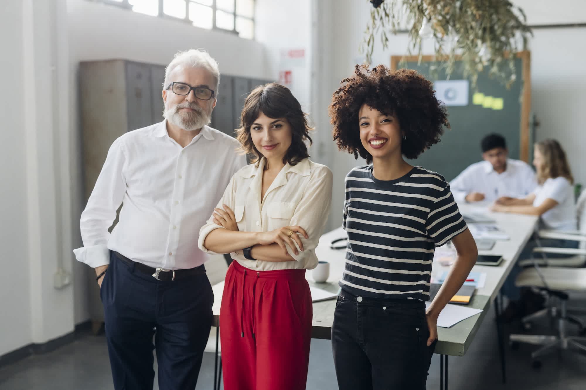Three people stand together and smile confidently in a modern office, with a man in a white shirt and two women, one in a white blouse and red pants and the other in a striped shirt; coworkers are working in the background.