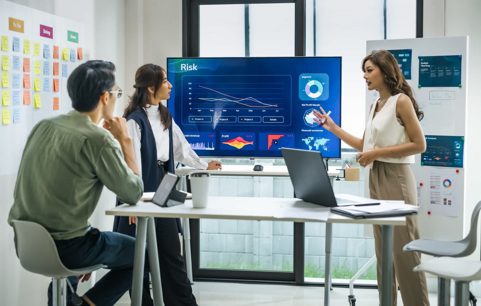 Three professionals in a modern office discuss risk analysis, with one woman presenting charts on a large screen while two colleagues listen. Laptops, notes, and a whiteboard are visible in the workspace.