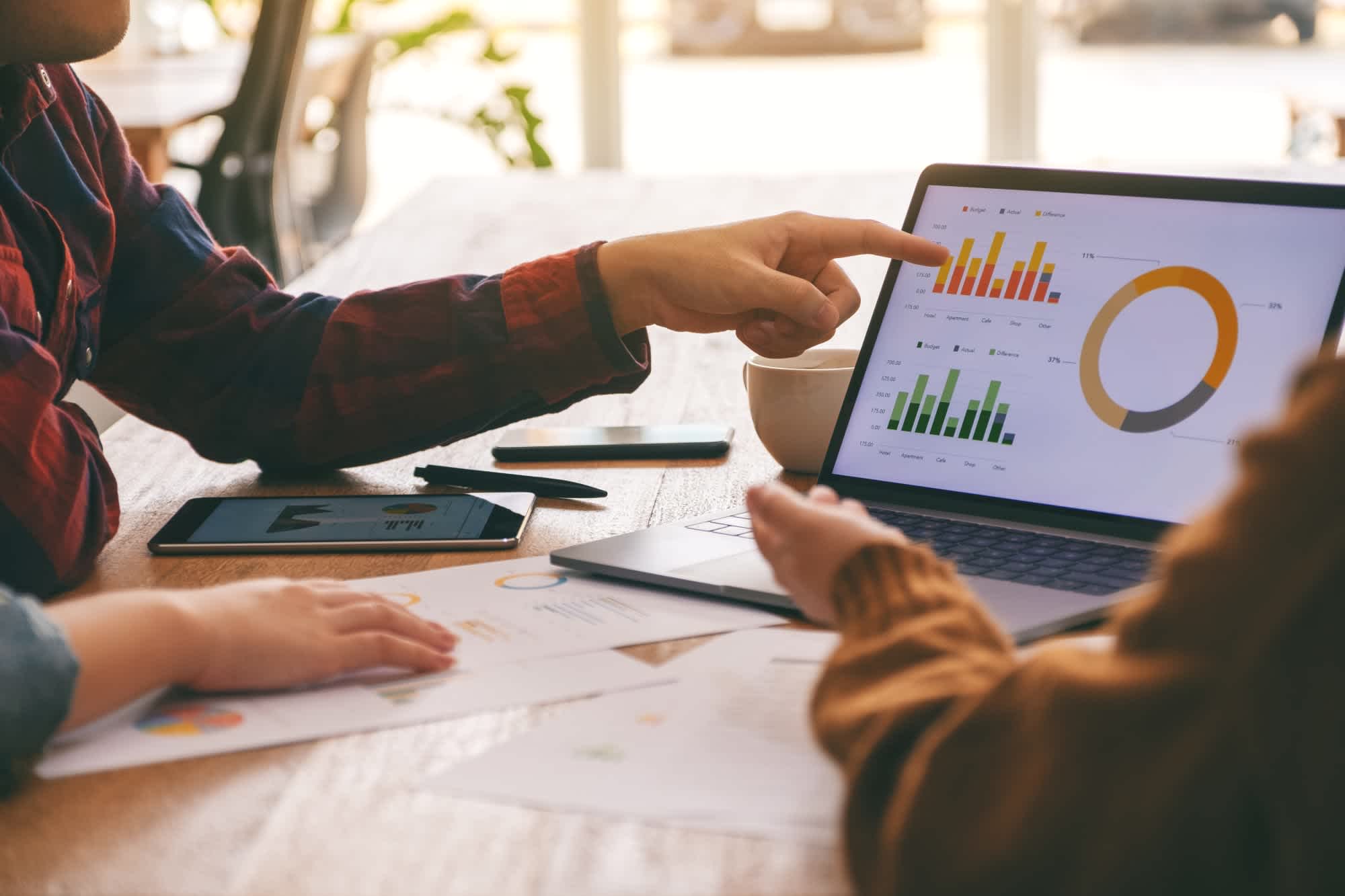 Two people at a table review charts and graphs on a laptop. One person points at the screen, which displays colorful bar and pie charts. Papers and a tablet are also on the table.