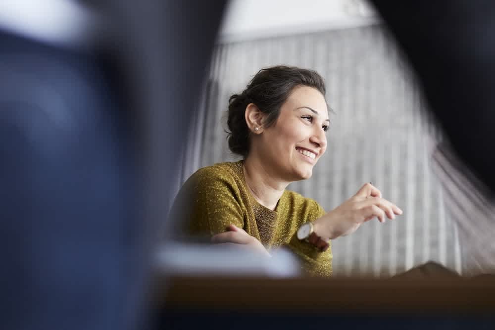 A woman with dark hair tied back, wearing a mustard yellow sweater and a watch, is smiling and gesturing with her hand while sitting indoors, with a blurred background and foreground.