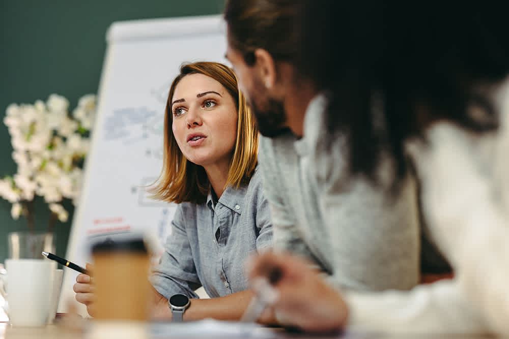 A woman speaks during a meeting, sitting at a table with two colleagues. A flip chart with diagrams is behind her, and there are flowers in the background. She holds a pen and appears engaged in conversation.