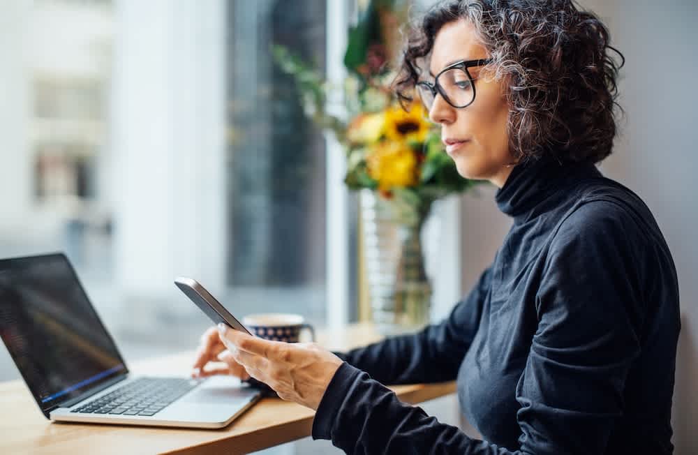 A woman with curly hair and glasses sits by a window, working at a laptop and holding a smartphone. A coffee mug and a vase with yellow flowers are on the table beside her.