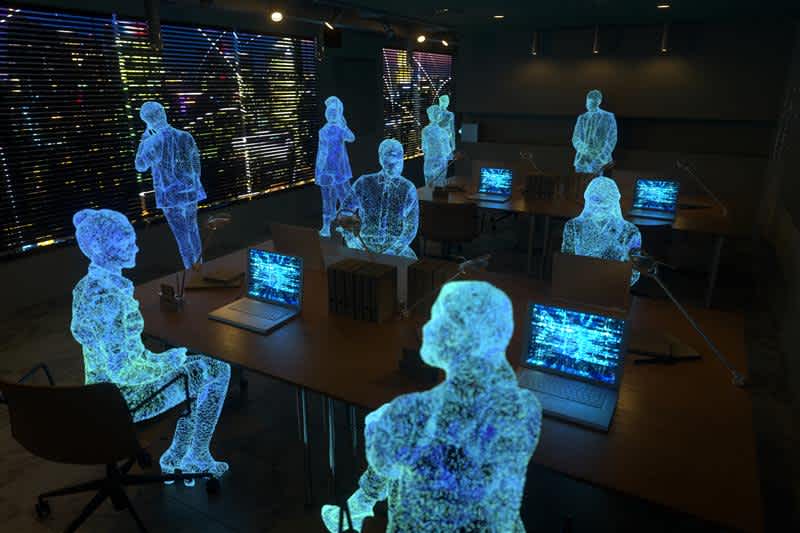 A dimly lit office with desks, laptops, and digital hologram figures of people sitting and standing, suggesting a futuristic or virtual work environment. Blinds cover the windows, revealing city lights outside.