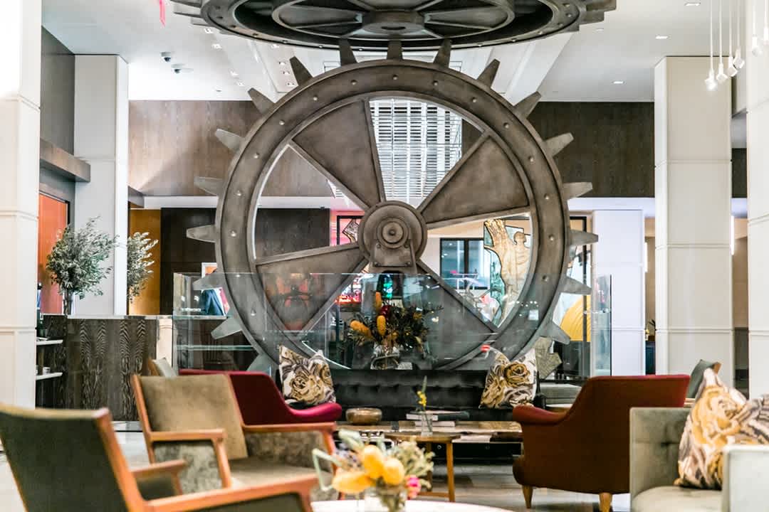 A hotel lobby features cozy seating with colorful armchairs and couches, decorative pillows, flowers, and a large industrial gear sculpture as the central focal point.