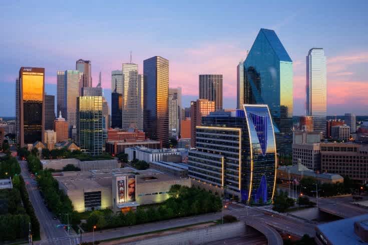 Downtown Dallas skyline at sunset, featuring modern and glass skyscrapers reflecting light, with highways and green trees in the foreground.