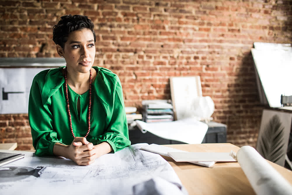 A woman in a green blouse and beaded necklace leans on a desk covered with architectural plans, looking thoughtfully to the side in a modern office with an exposed brick wall.