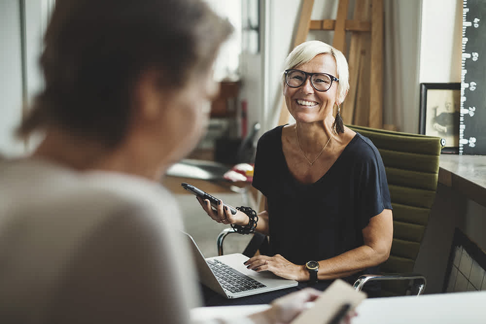 Smiling woman with short white hair and glasses holds a smartphone and sits at a desk with a laptop, facing another person in an office setting.