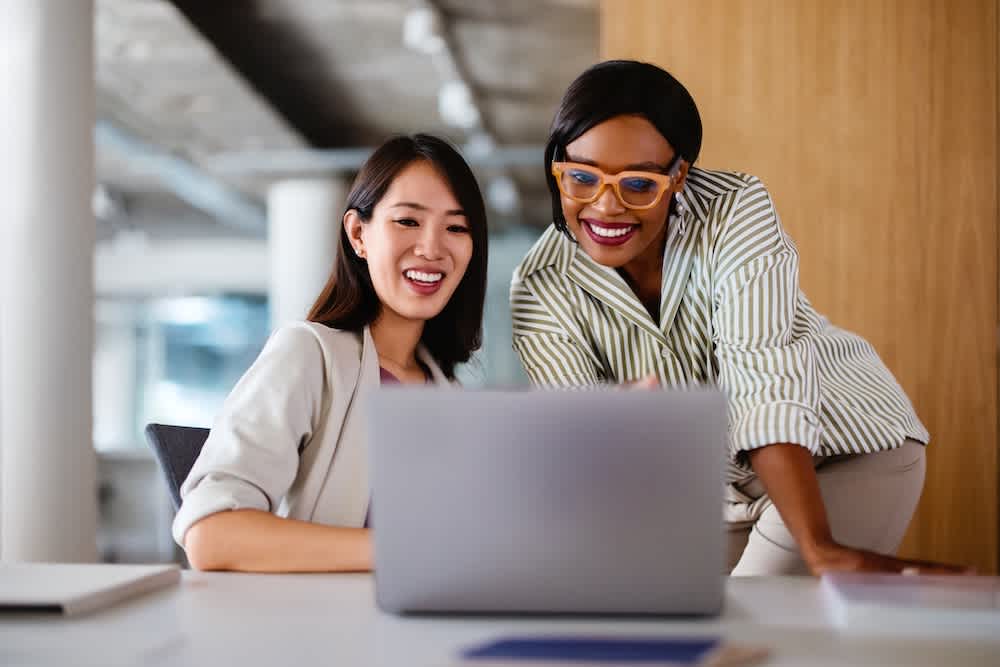 Two women in business attire smiling and looking at a laptop together in a modern office setting. One is seated, and the other is standing, wearing glasses and leaning in.