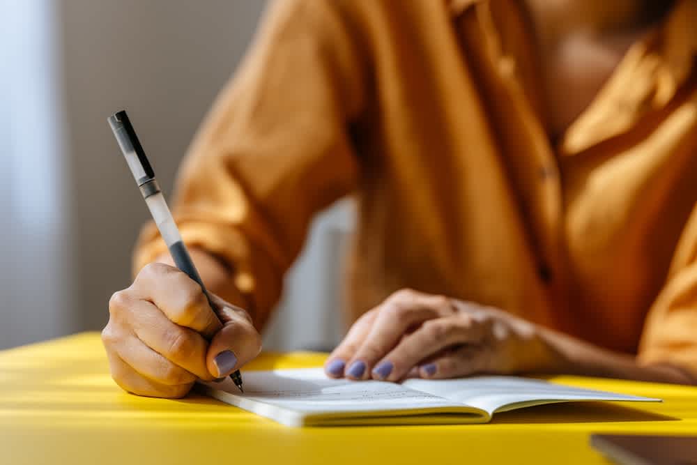 Close-up of a person wearing an orange shirt, holding a pen, and writing in a notebook on a yellow table. Only the hands and part of the torso are visible.