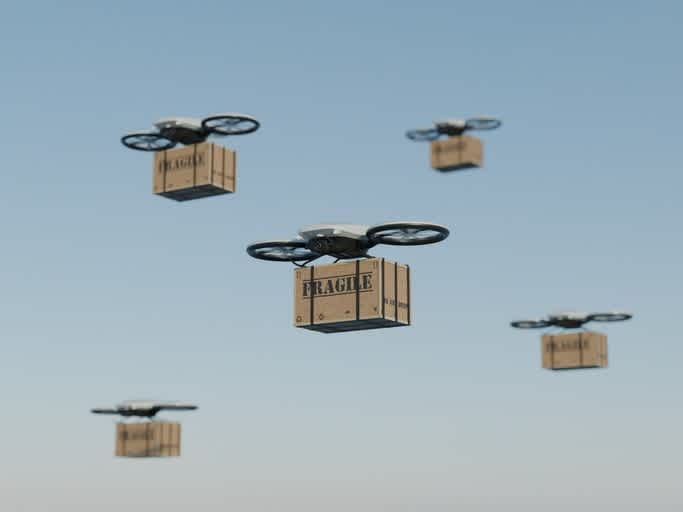 Several drones fly in a clear blue sky, each carrying a box labeled FRAGILE underneath, demonstrating a concept of drone delivery for fragile packages.