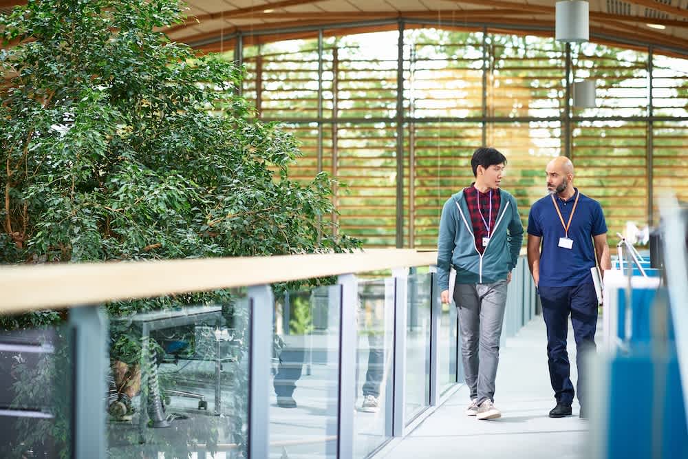 Two men walk side by side in a modern building with large windows and plants, engaged in conversation. Both wear lanyards, and sunlight streams through the wooden slats behind them.