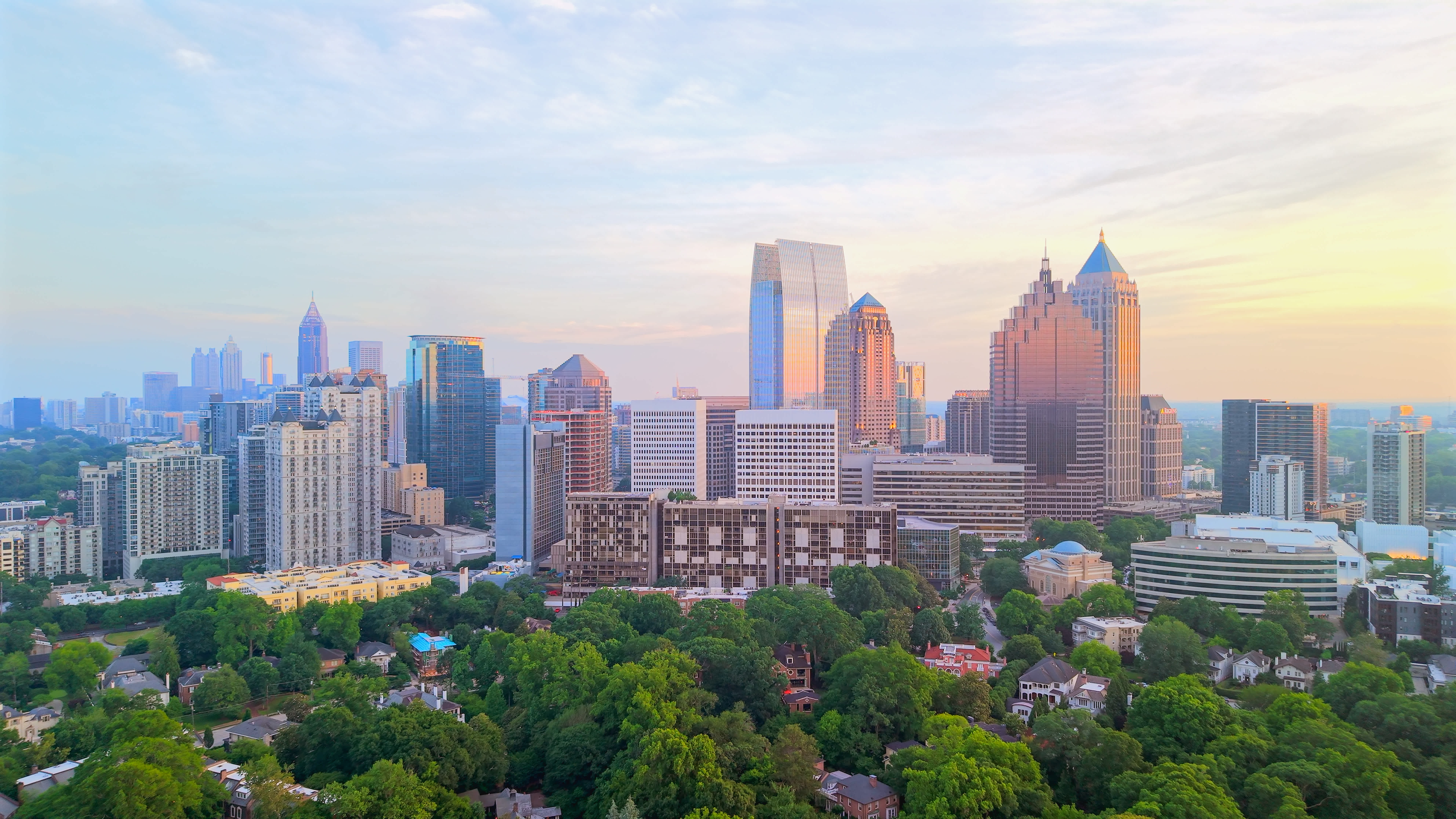 A cityscape of Atlanta, Georgia, shows modern skyscrapers and office buildings with lush green trees in the foreground under a pastel-colored sky at sunset.