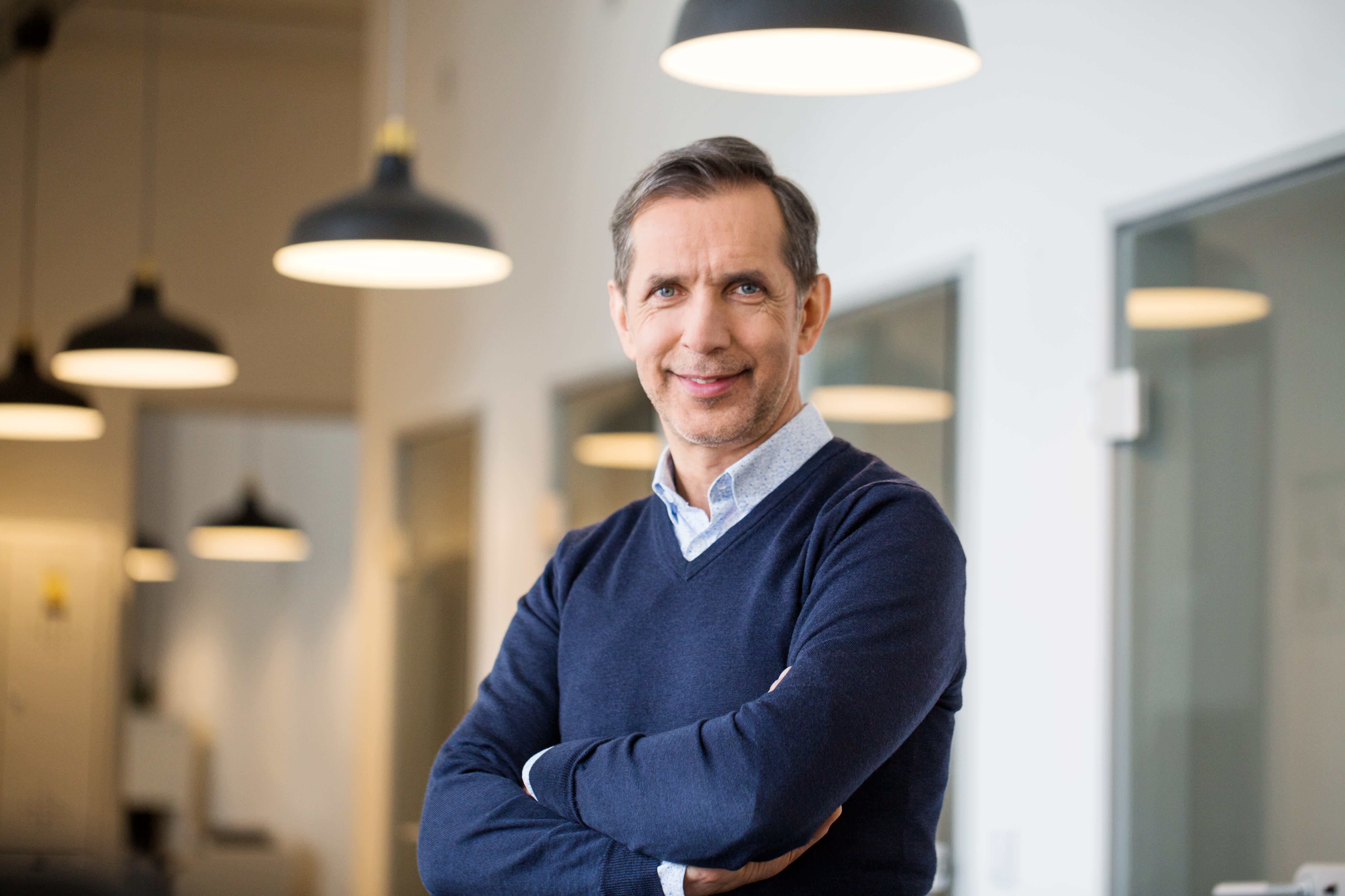 A middle-aged man with short brown hair, wearing a navy blue sweater over a collared shirt, stands with arms crossed and smiles in a modern office with hanging lights and glass doors.