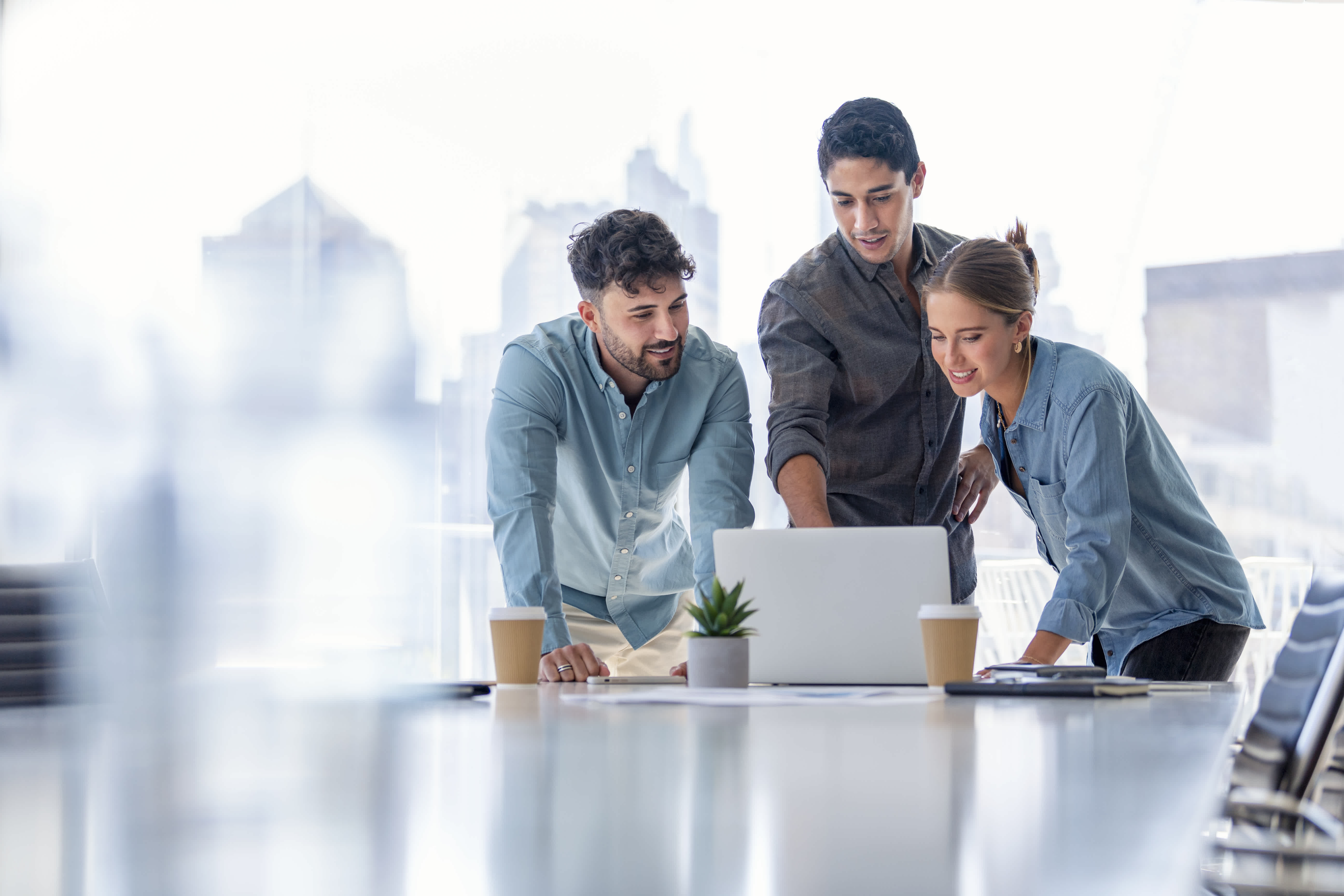 Three colleagues stand around a table looking at a laptop screen, discussing work in a bright modern office with large windows and city views. Two coffee cups and a small plant are on the table.