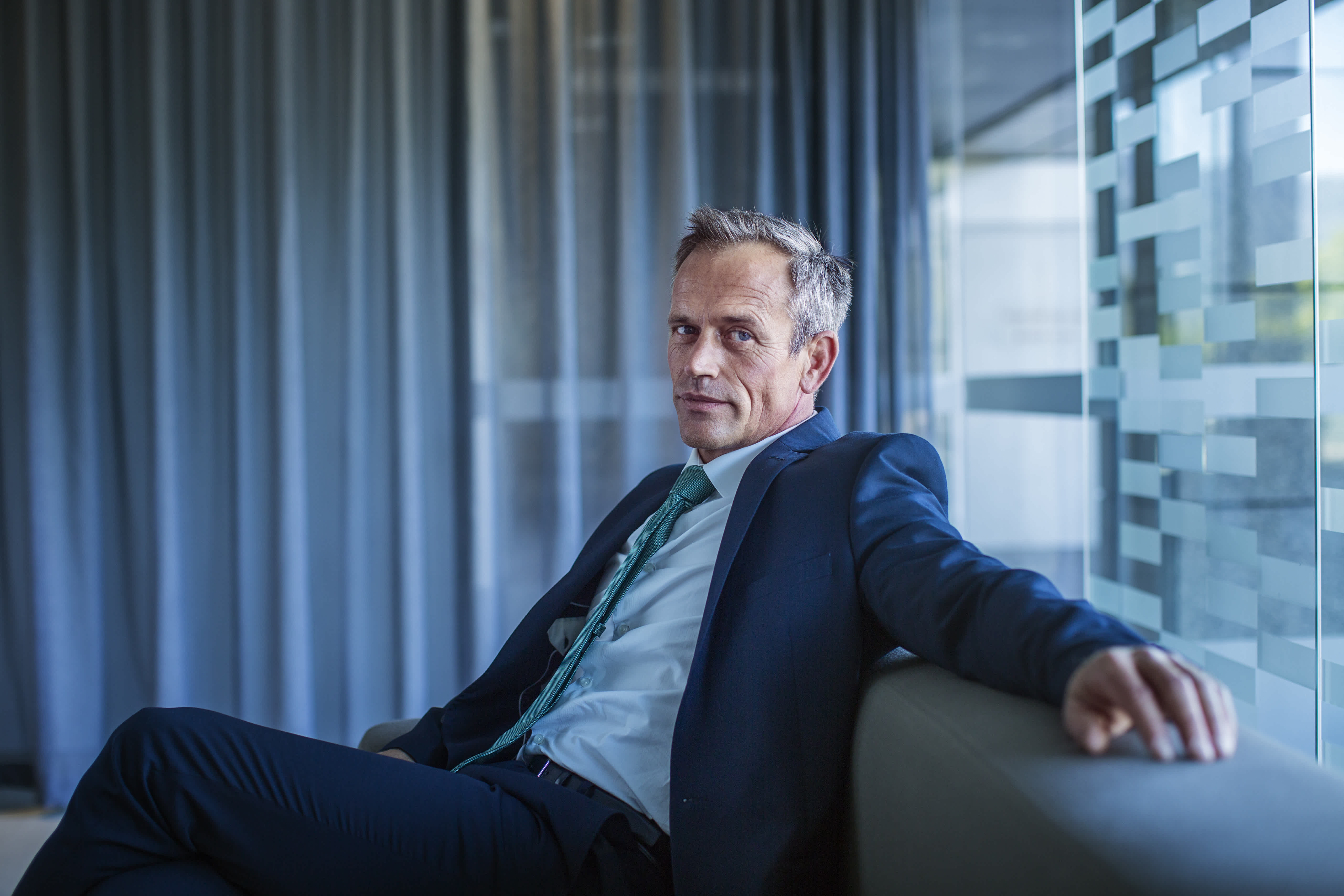 A middle-aged man in a suit sits on a sofa in a modern office, looking confidently at the camera. He has short gray hair and is relaxed, with one arm resting on the back of the sofa near a glass wall.