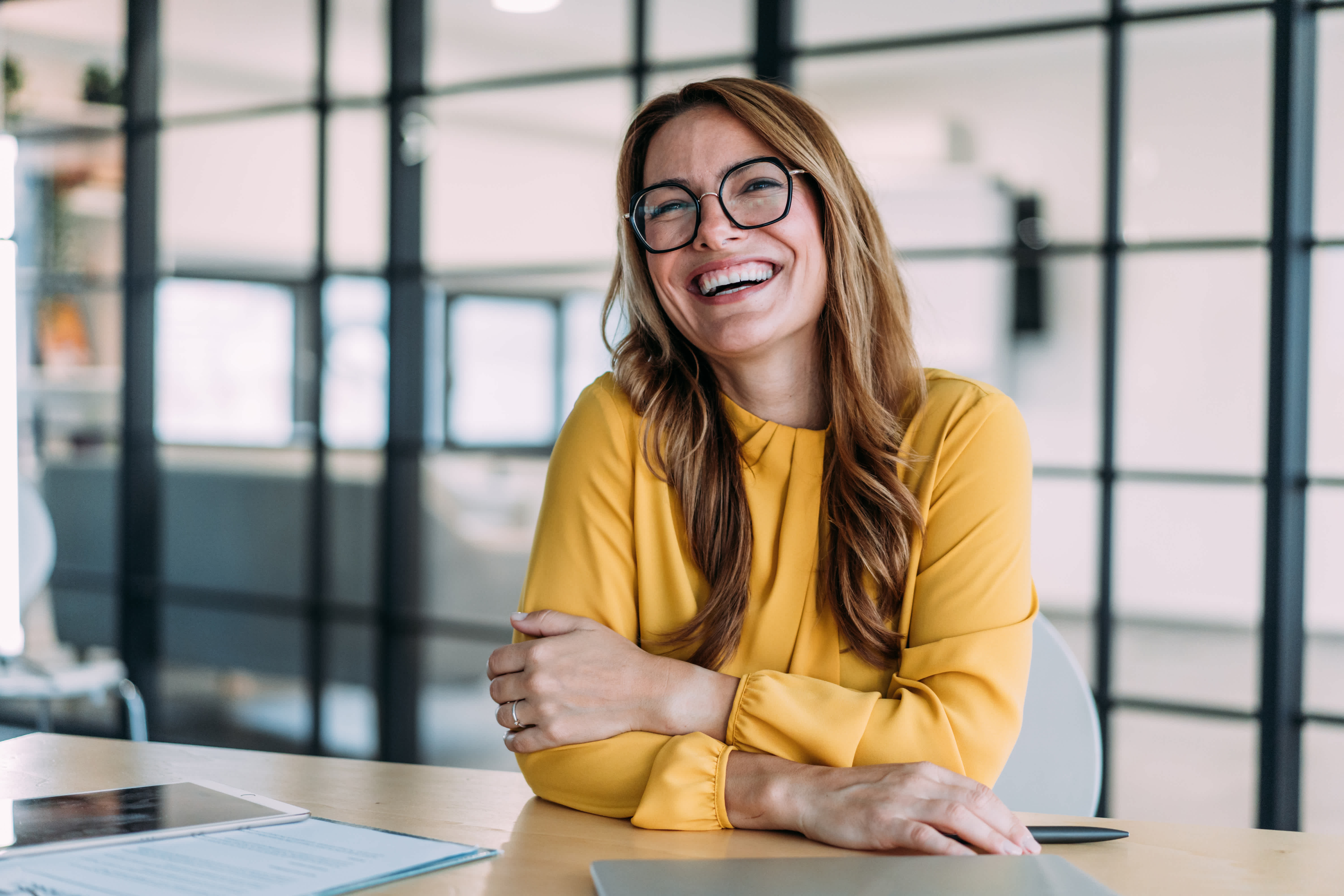 A woman with long brown hair and glasses, wearing a yellow blouse, sits at a desk smiling and laughing in a modern office with glass walls.
