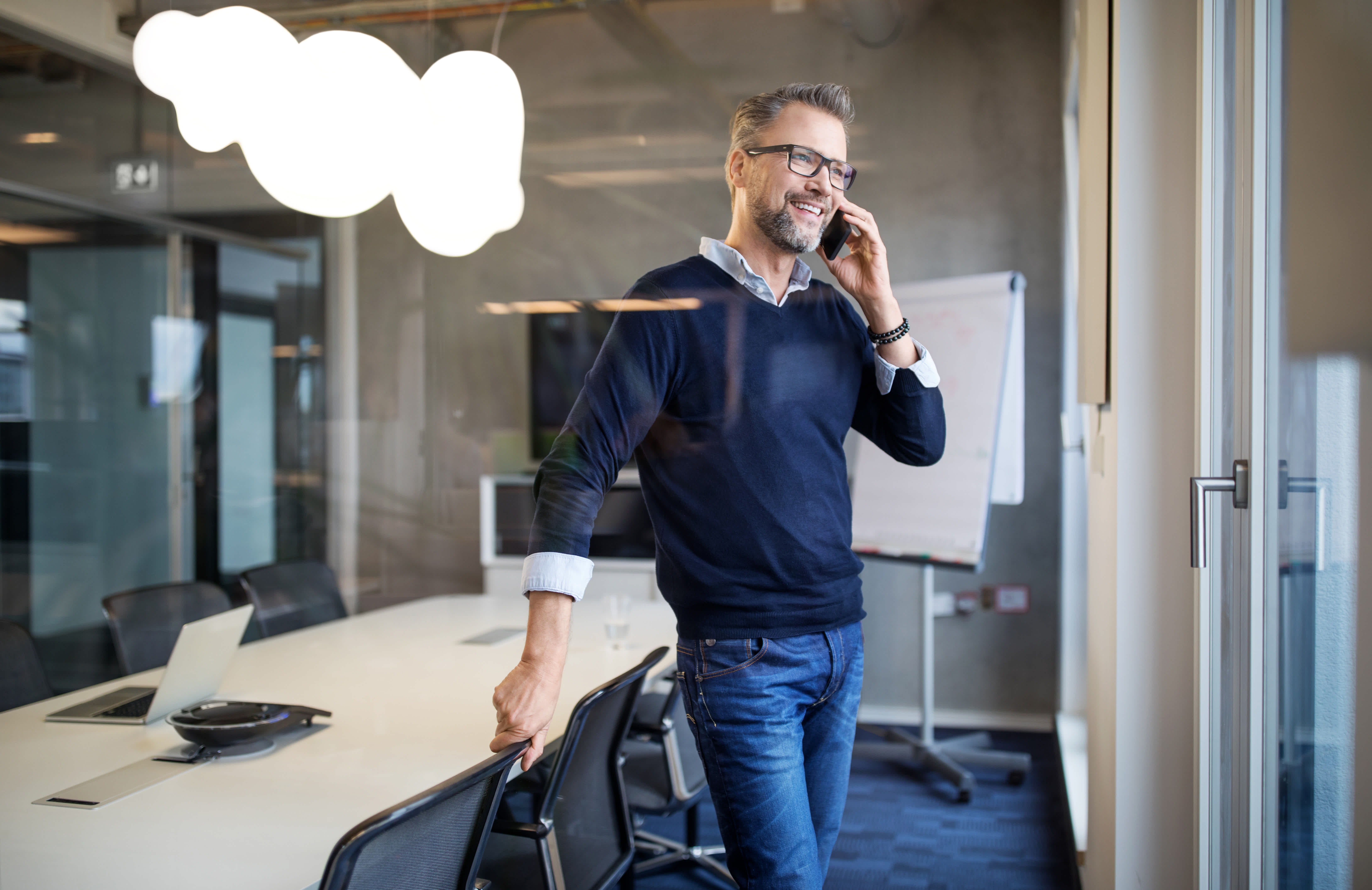 A man wearing glasses, a navy sweater, and jeans smiles while talking on a smartphone in a modern conference room with a large table, chairs, and a whiteboard.