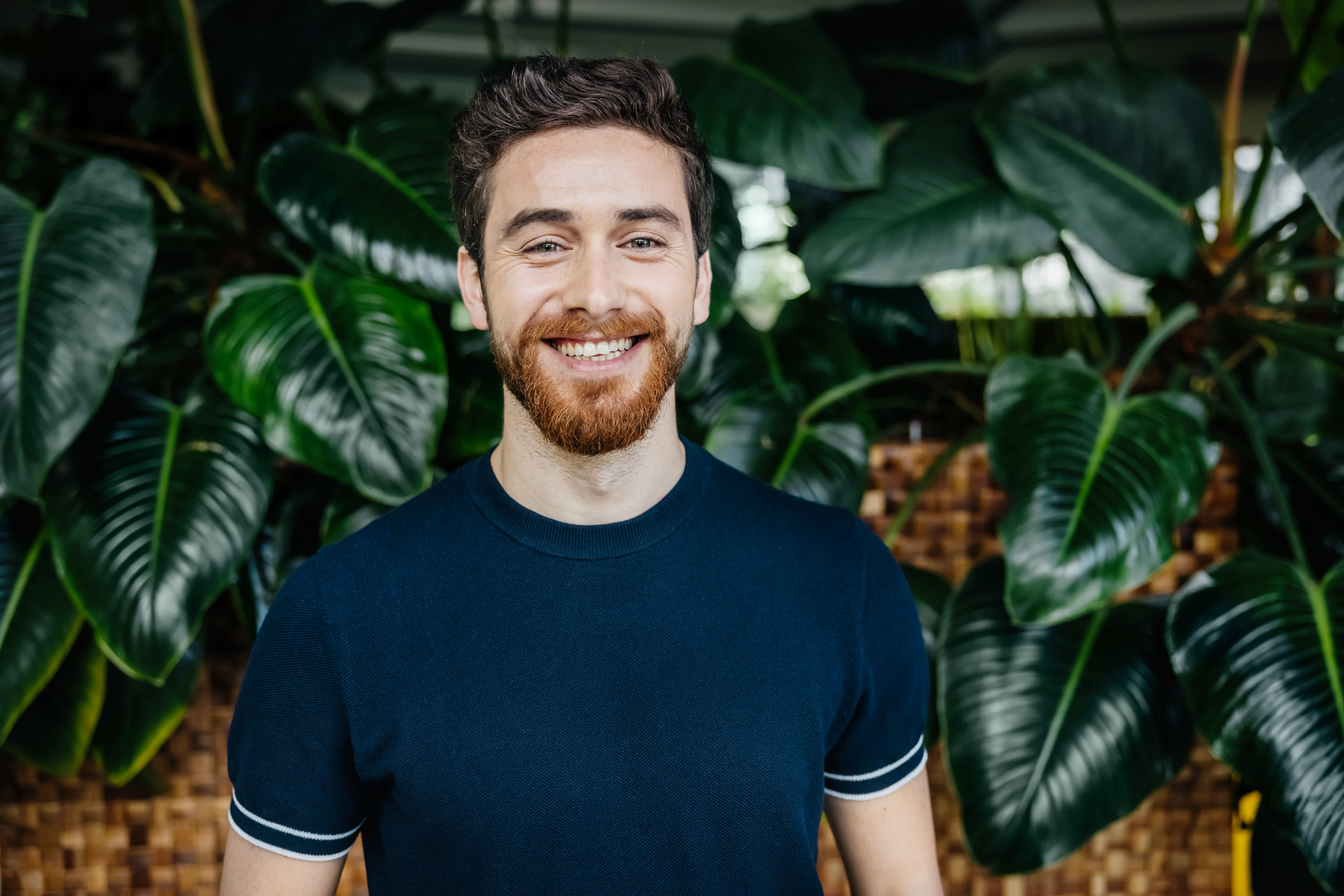 A man with a beard and mustache, wearing a navy blue shirt, smiles while standing in front of large green leafy plants.
