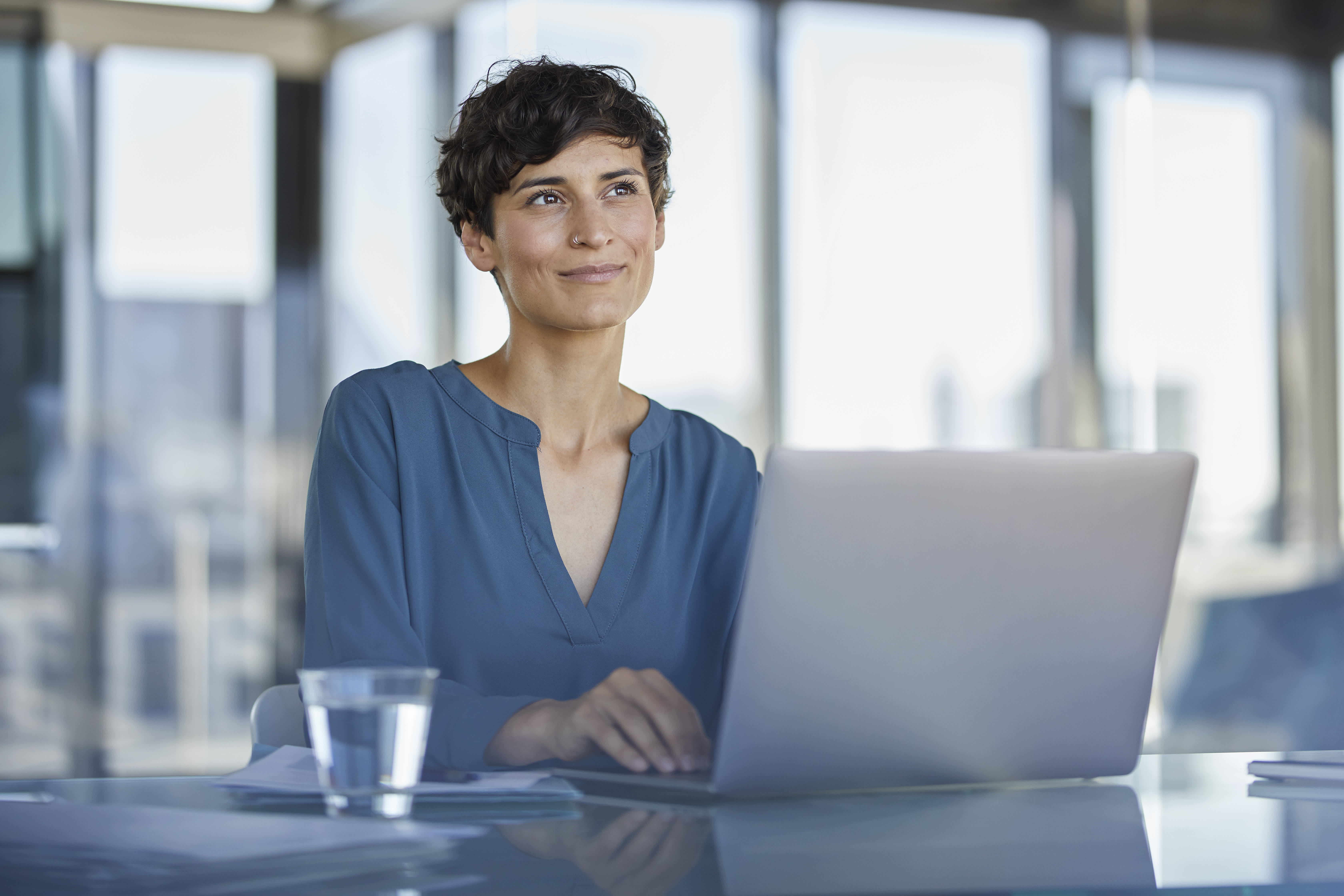 A person with short hair sits at a desk in an office, smiling slightly while looking away from their laptop. There is a glass of water and some papers on the desk. Large windows provide natural light.