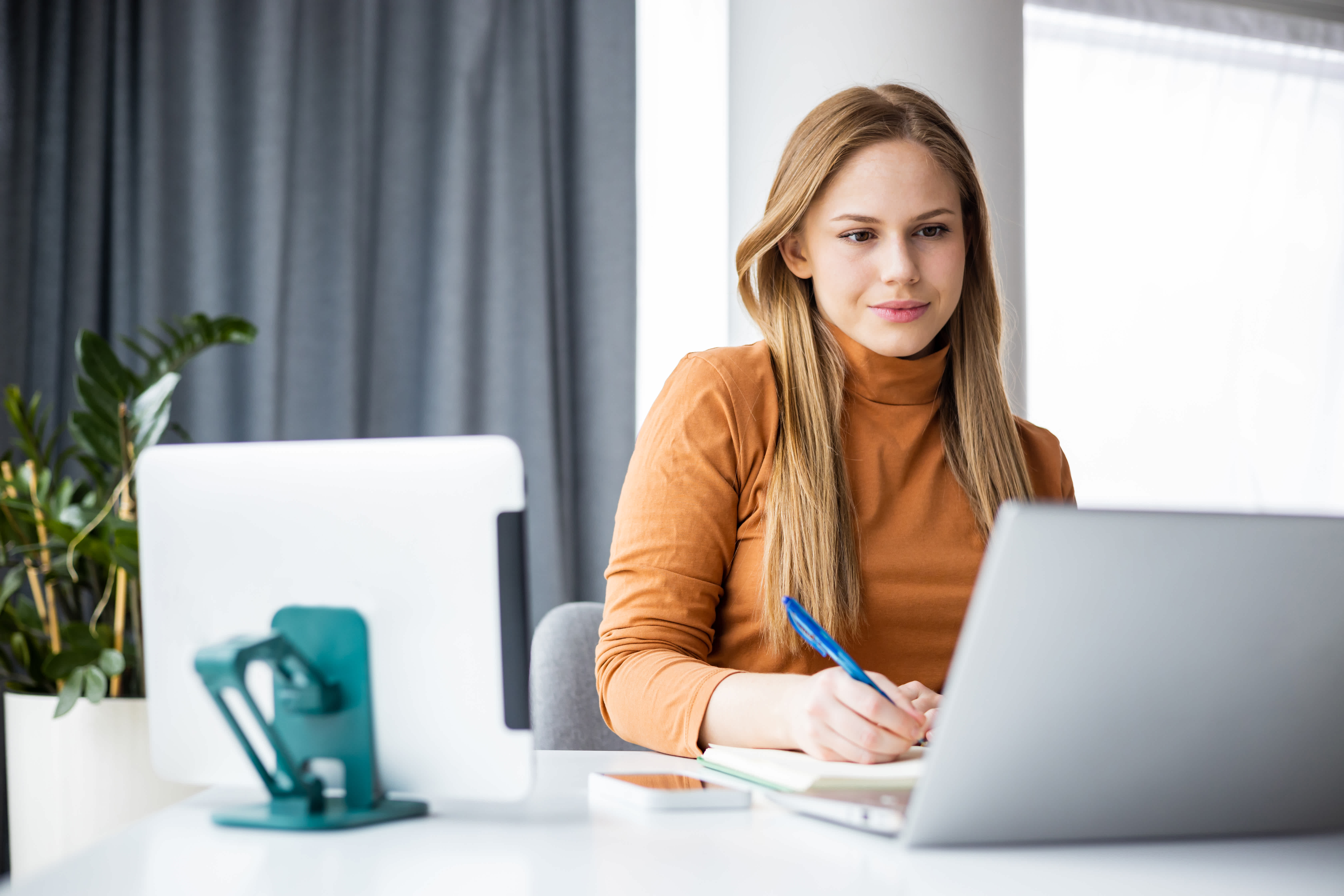 A young woman sits at a desk, looking at a laptop and writing in a notebook with a pen. She appears focused, with a tablet, phone holder, and potted plant visible on the desk in a bright, modern room.
