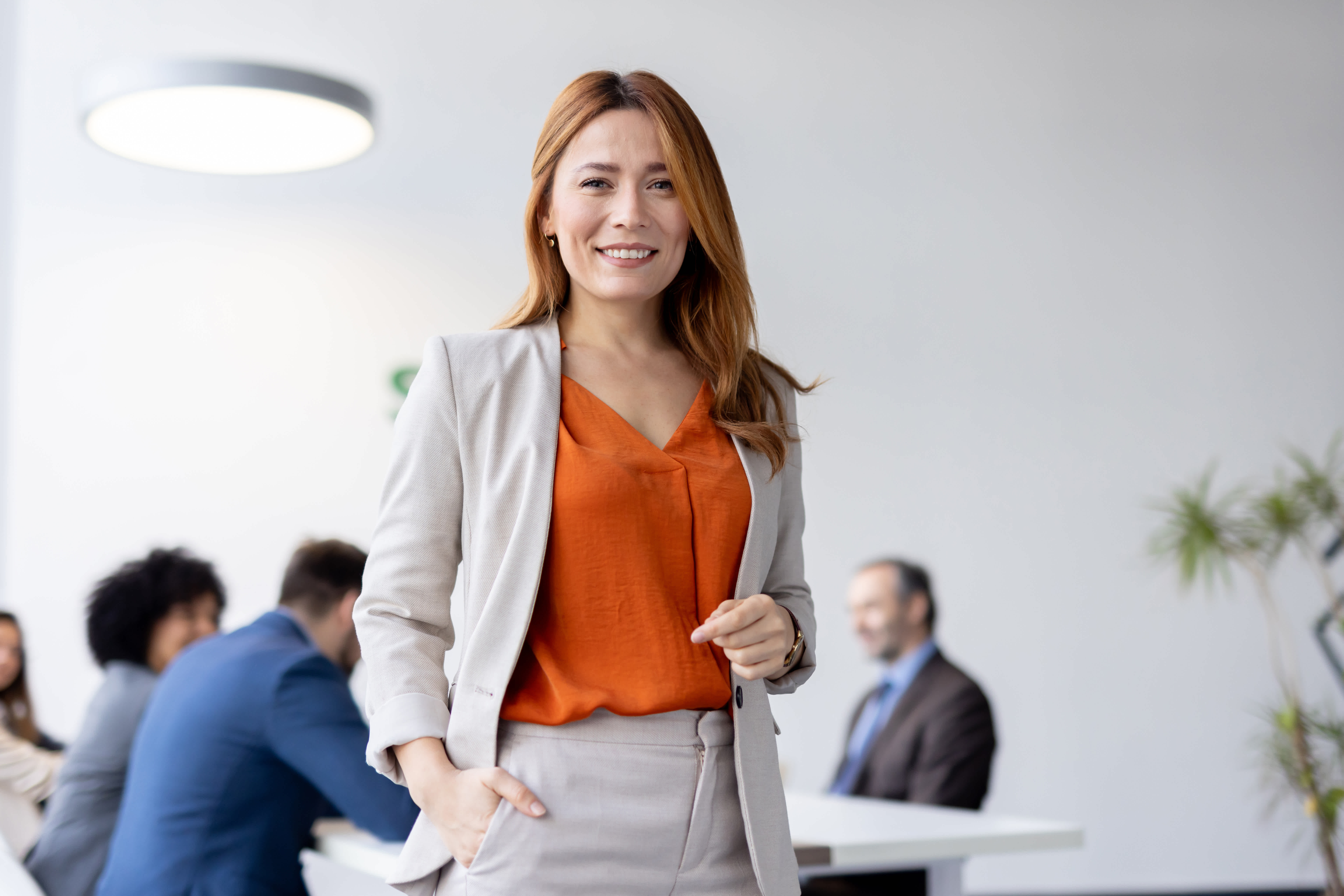 A confident woman with long reddish-brown hair, wearing a beige suit and orange blouse, stands smiling in a modern office. Several people are blurred in the background, engaged in a meeting around a table.